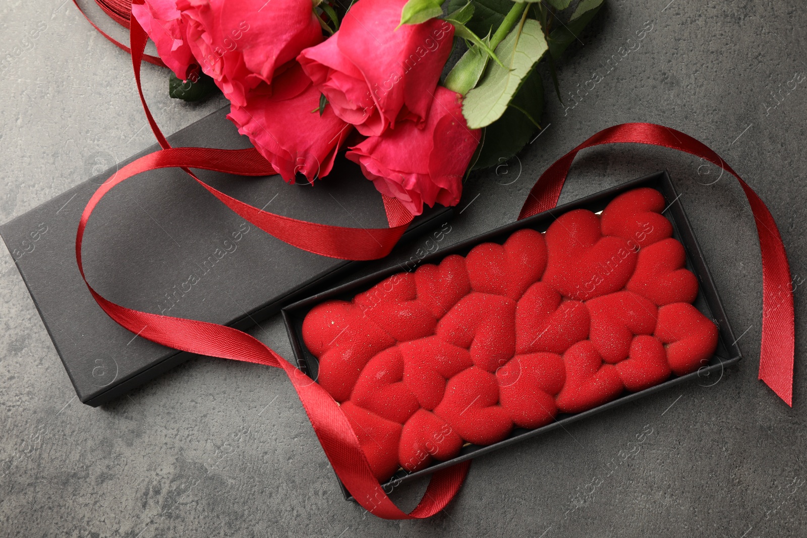 Photo of Chocolate bar decorated with hearts in box, roses and red ribbon on grey table, flat lay