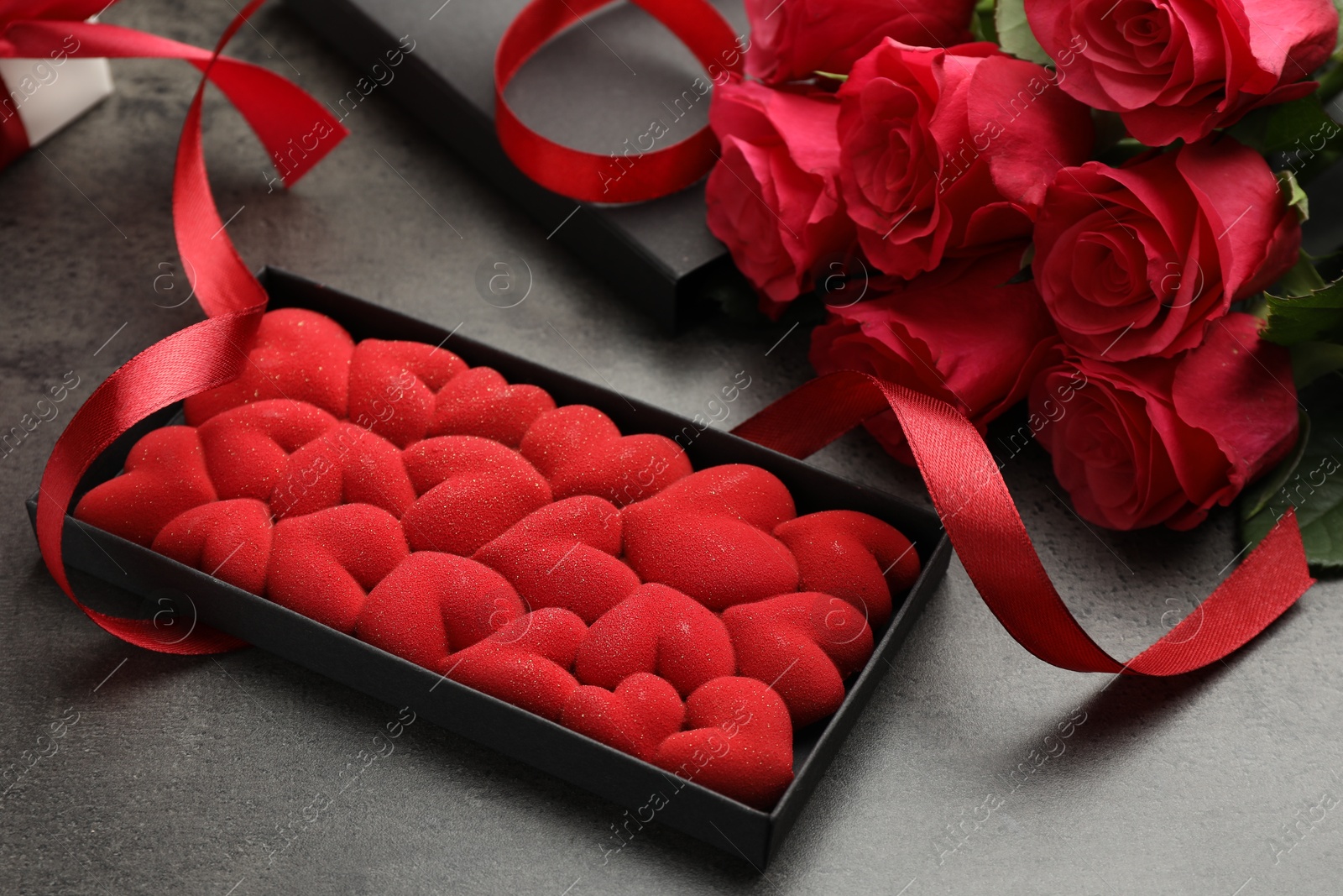 Photo of Chocolate bar decorated with hearts in box, roses and red ribbon on grey table, closeup