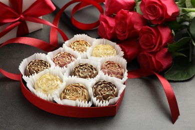 Photo of Flower shaped chocolate bonbons in box, red roses, gift and ribbon on grey table, closeup