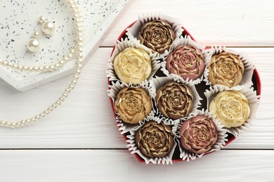 Photo of Flower shaped chocolate bonbons in box and pearl jewelry on white wooden table, flat lay