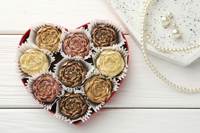 Photo of Flower shaped chocolate bonbons in box and pearl jewelry on white wooden table, flat lay