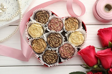 Photo of Flower shaped chocolate bonbons in box, red roses, pink ribbon and pearl jewelry on white wooden table, flat lay