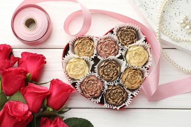 Photo of Flower shaped chocolate bonbons in box, red roses, pink ribbon and pearl jewelry on white wooden table, flat lay