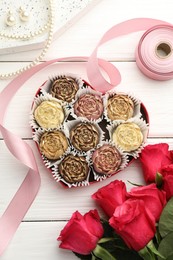 Photo of Flower shaped chocolate bonbons in box, red roses, pink ribbon and pearl jewelry on white wooden table, flat lay