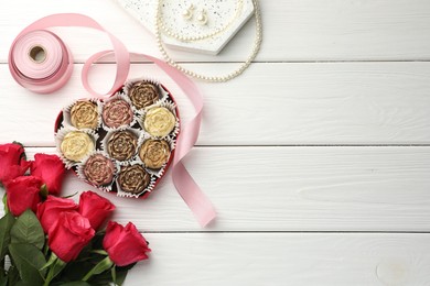 Photo of Flower shaped chocolate bonbons in box, red roses, pink ribbon and pearl jewelry on white wooden table, flat lay. Space for text