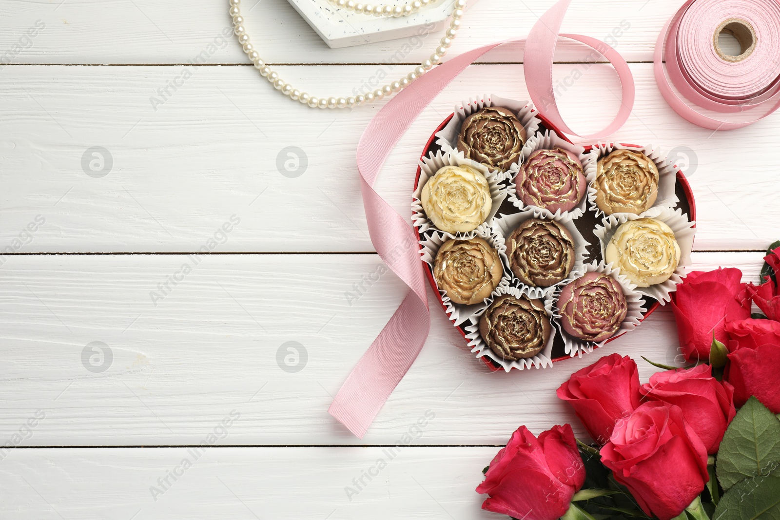 Photo of Flower shaped chocolate bonbons in box, red roses, pink ribbon and pearl jewelry on white wooden table, flat lay. Space for text