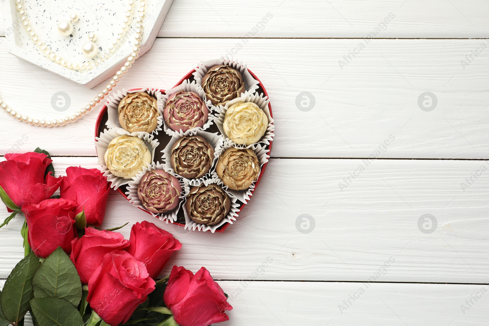 Photo of Flower shaped chocolate bonbons in box, red roses and pearl jewelry on white wooden table, flat lay. Space for text