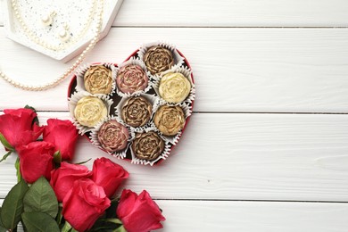 Photo of Flower shaped chocolate bonbons in box, red roses and pearl jewelry on white wooden table, flat lay. Space for text