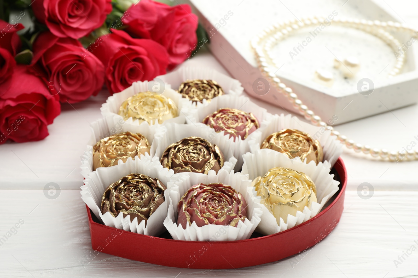 Photo of Flower shaped chocolate bonbons in box, red roses and pearl jewelry on white wooden table, closeup