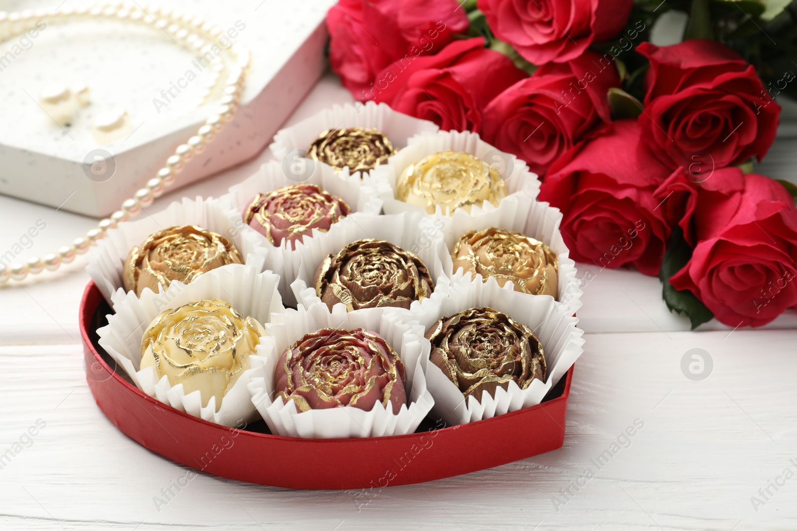 Photo of Flower shaped chocolate bonbons in box, red roses and pearl jewelry on white wooden table, closeup