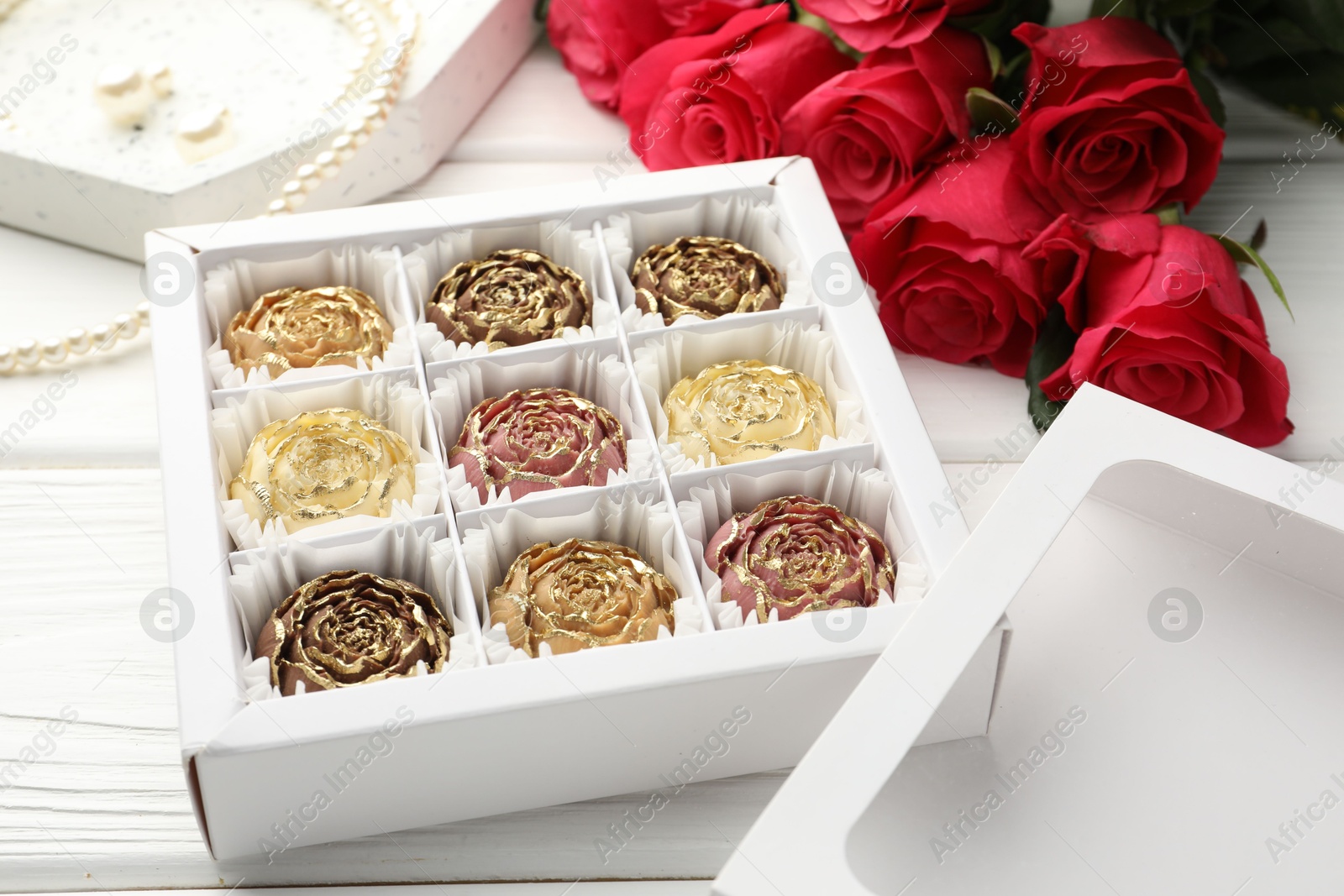 Photo of Flower shaped chocolate bonbons in box, red roses and pearl jewelry on white wooden table, closeup