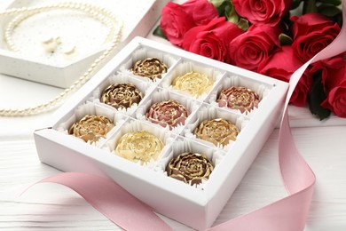 Photo of Flower shaped chocolate bonbons in box, red roses, pink ribbon and pearl jewelry on white wooden table, closeup