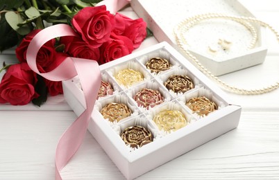 Photo of Flower shaped chocolate bonbons in box, red roses, pink ribbon and pearl jewelry on white wooden table, closeup