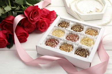 Photo of Flower shaped chocolate bonbons in box, red roses, pink ribbon and pearl jewelry on white wooden table, closeup