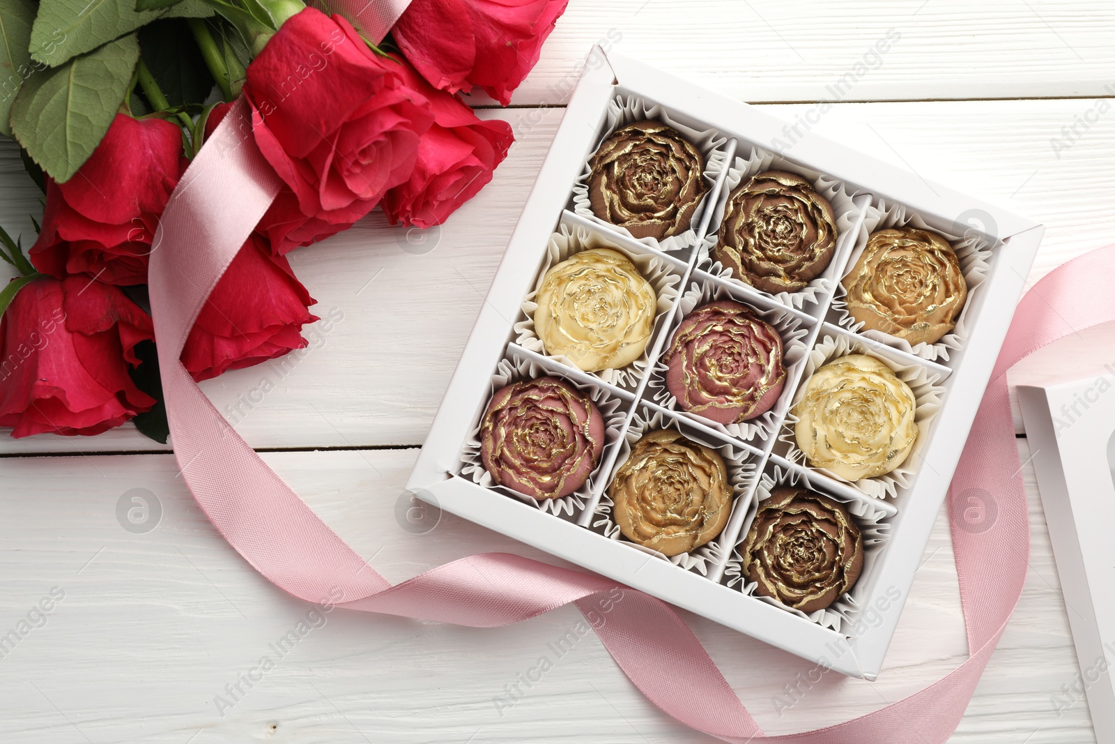 Photo of Flower shaped chocolate bonbons in box, red roses and pink ribbon on white wooden table, flat lay