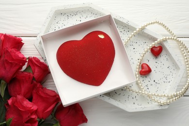 Photo of Heart shaped chocolate in box, red roses and pearl jewelry on white wooden table, flat lay