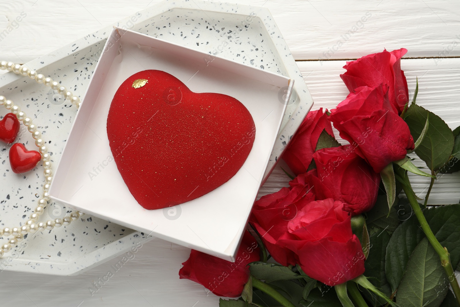 Photo of Heart shaped chocolate in box, red roses and pearl jewelry on white wooden table, flat lay