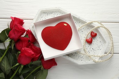 Photo of Heart shaped chocolate in box, red roses and pearl jewelry on white wooden table, flat lay