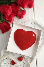 Photo of Heart shaped chocolate in box, red roses and pearl jewelry on white wooden table, flat lay