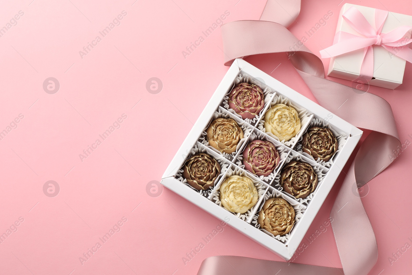 Photo of Flower shaped chocolate bonbons in box, gift and ribbon on pink background, flat lay. Space for text