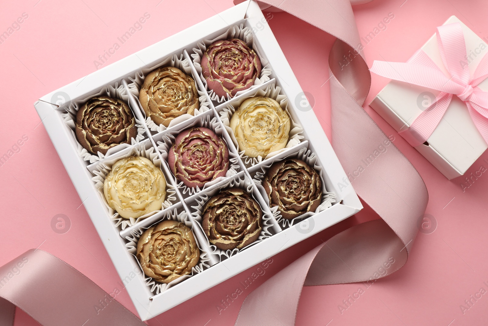 Photo of Flower shaped chocolate bonbons in box, gift and ribbon on pink background, flat lay