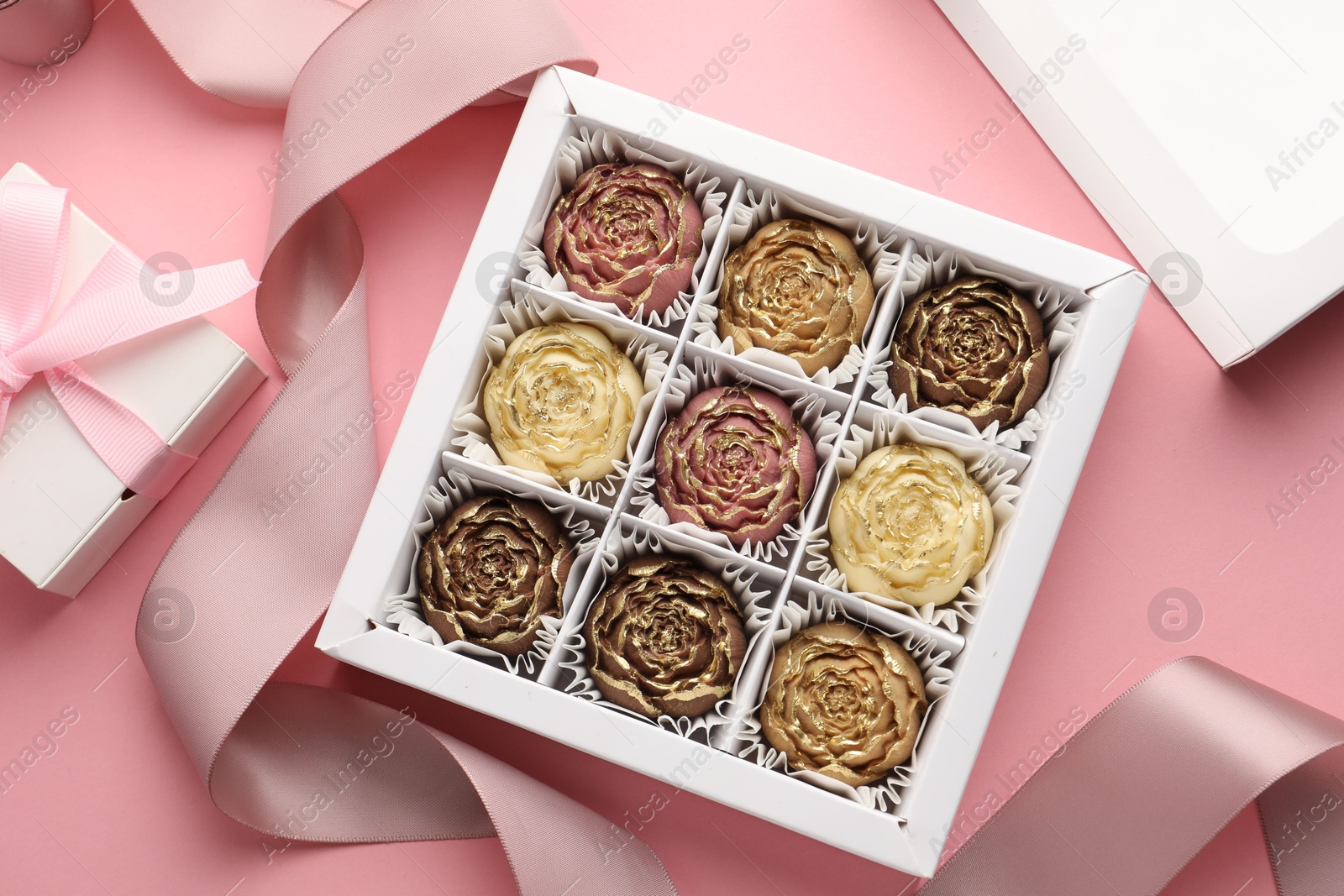 Photo of Flower shaped chocolate bonbons in box, gift and ribbon on pink background, flat lay