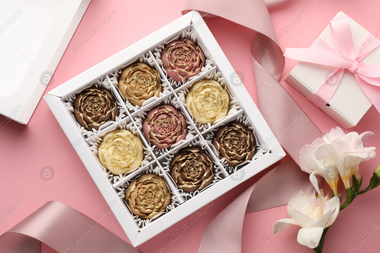 Photo of Flower shaped chocolate bonbons in box, gift, flowers and ribbon on pink background, flat lay