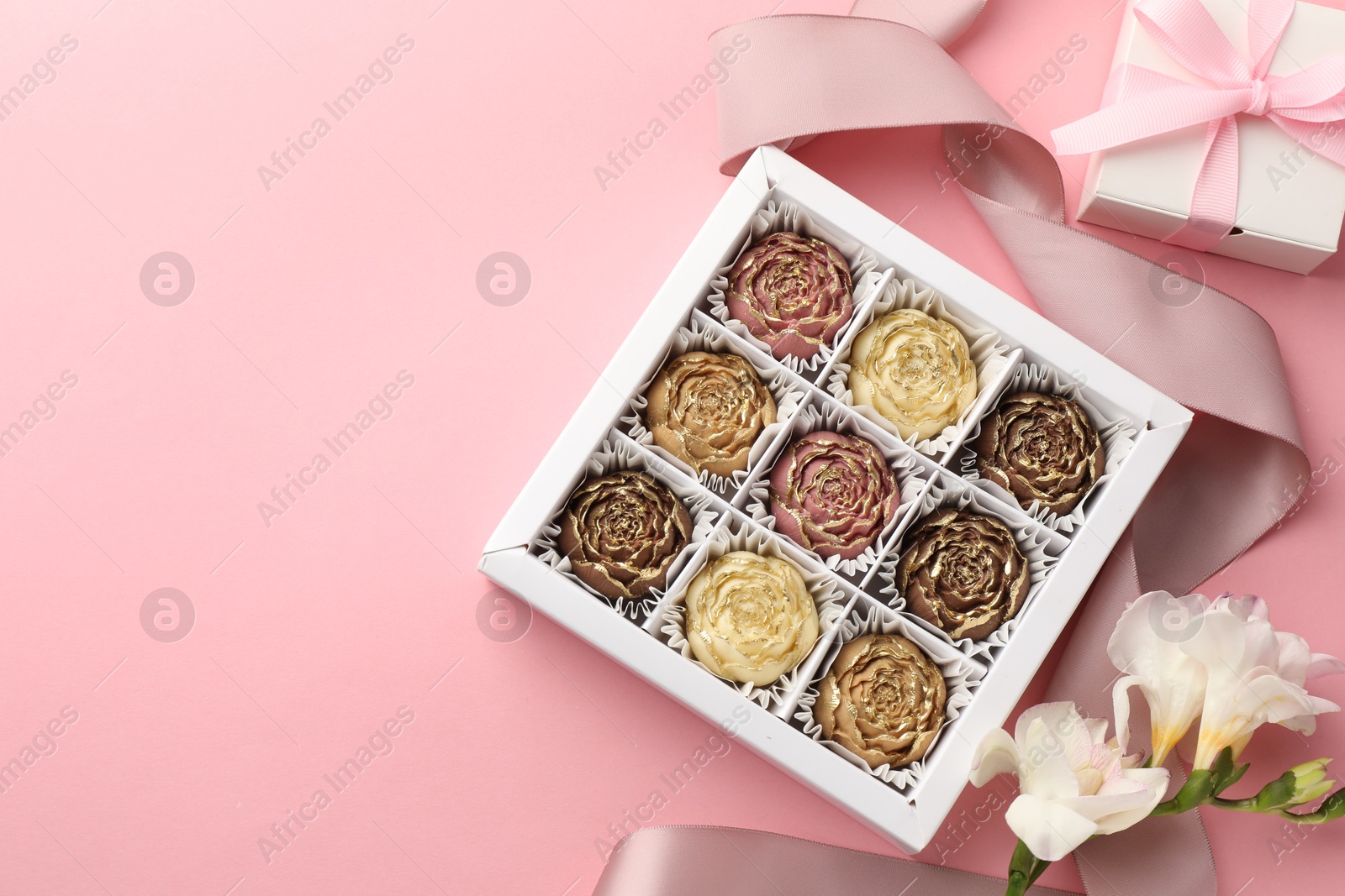 Photo of Flower shaped chocolate bonbons in box, gift, flowers and ribbon on pink background, flat lay. Space for text