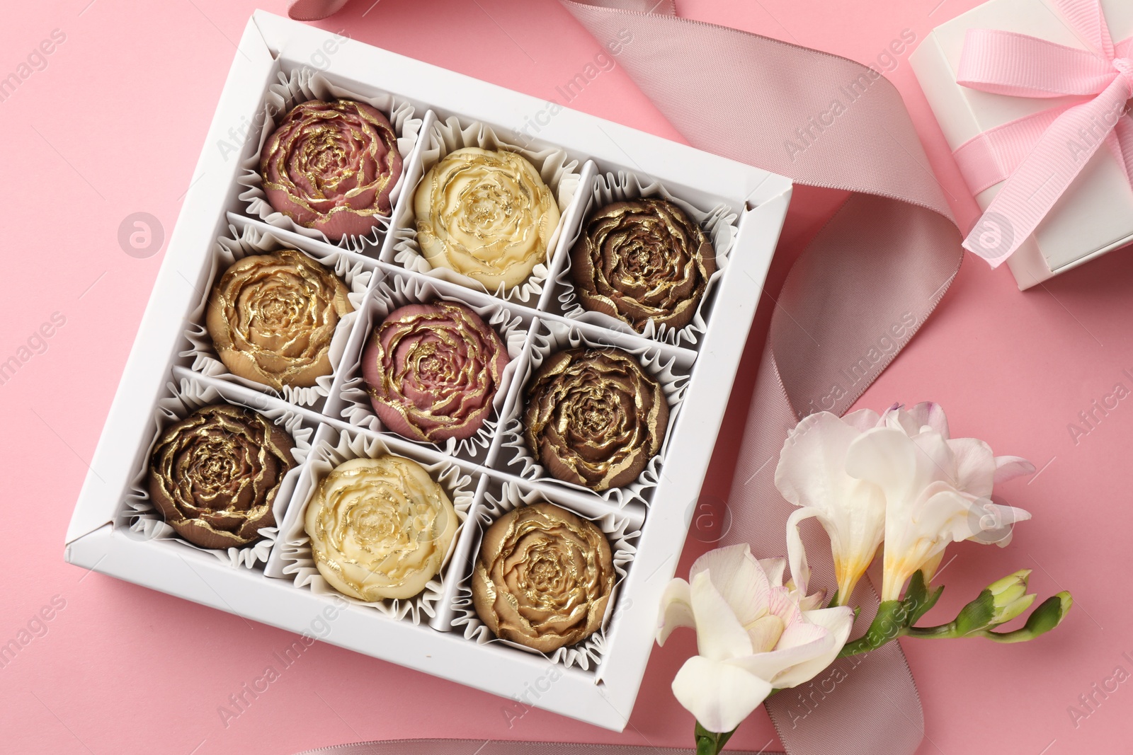 Photo of Flower shaped chocolate bonbons in box, gift, flowers and ribbon on pink background, flat lay