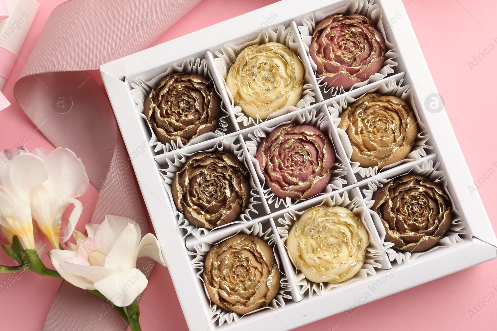 Photo of Flower shaped chocolate bonbons in box, flowers and ribbon on pink background, flat lay