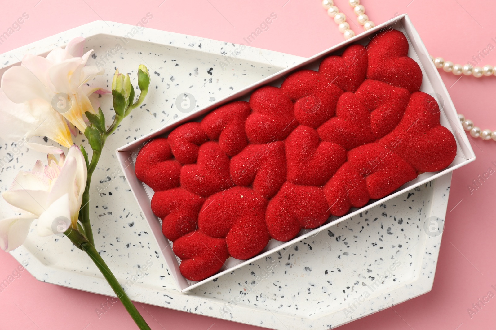 Photo of Chocolate bar decorated with hearts in box, flowers and pearl jewelry on pink background, flat lay