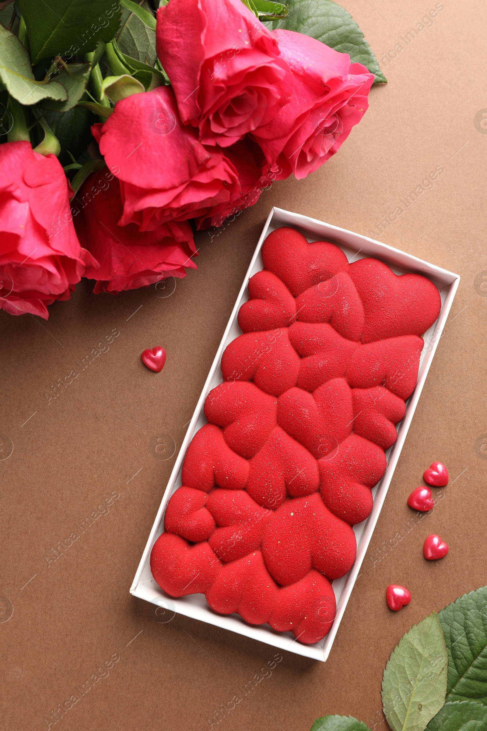 Photo of Chocolate bar decorated with hearts, confetti and roses on brown background, flat lay