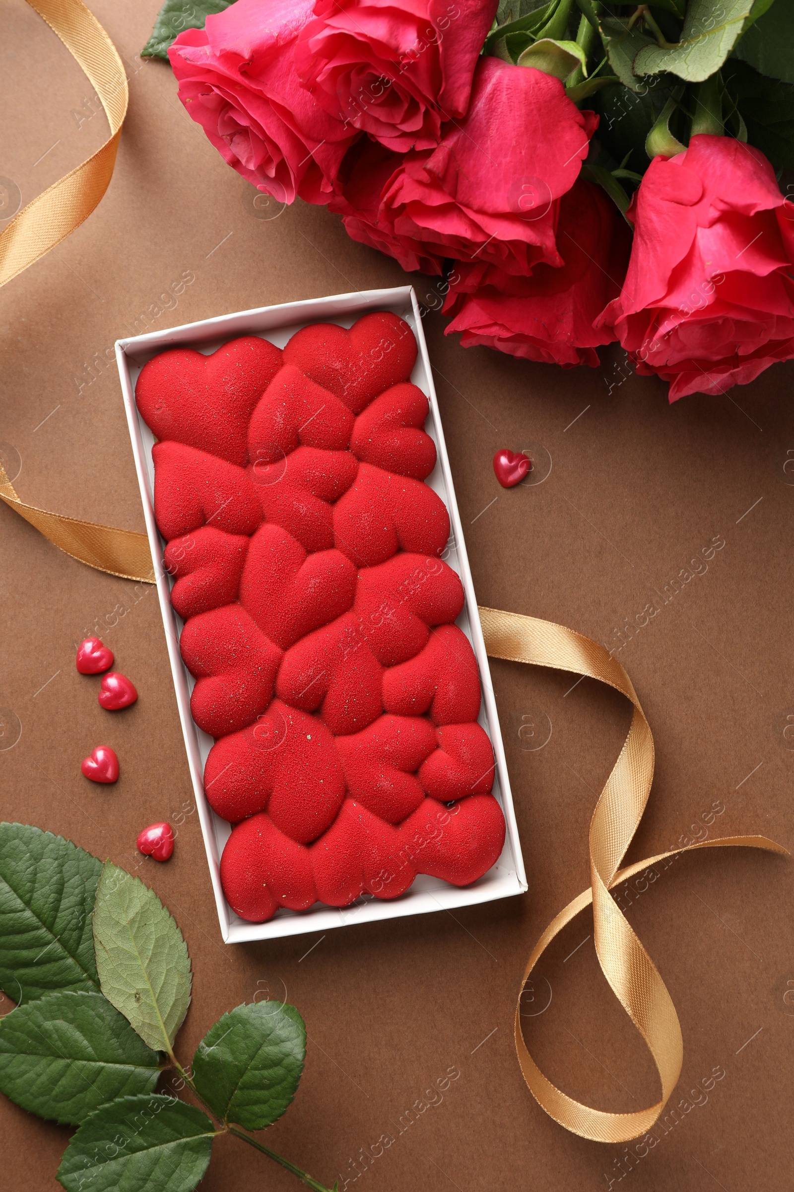 Photo of Chocolate bar decorated with hearts, confetti and roses on brown background, flat lay