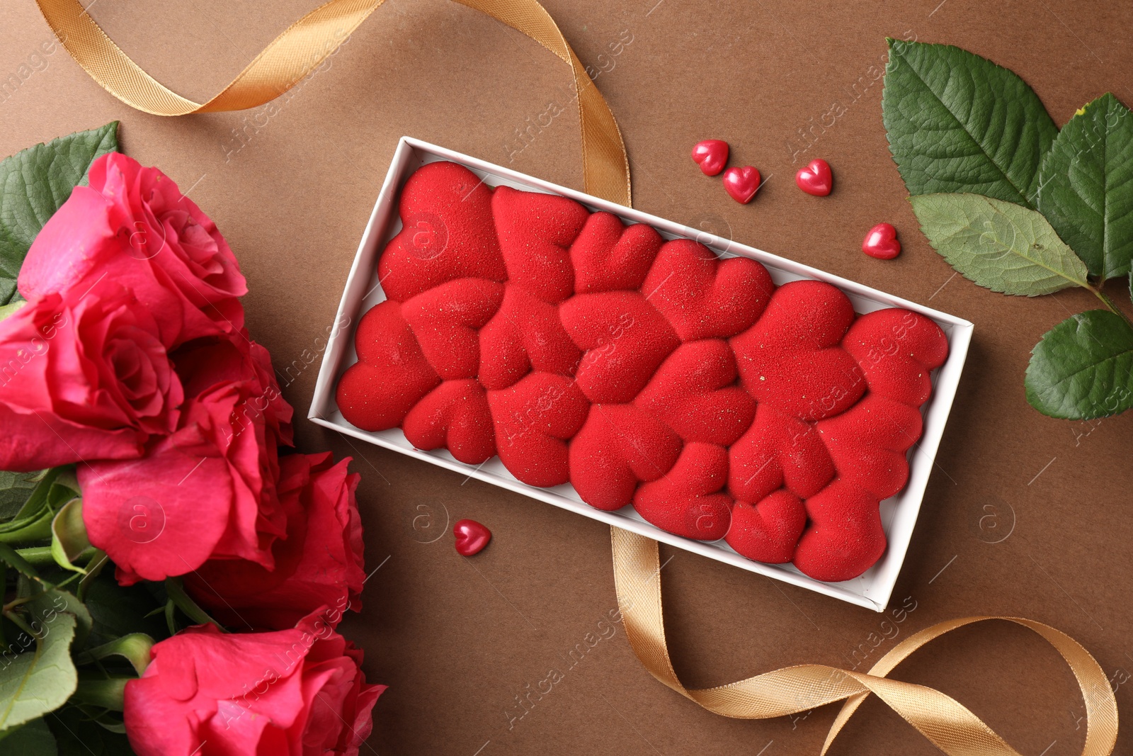 Photo of Chocolate bar decorated with hearts, confetti and roses on brown background, flat lay