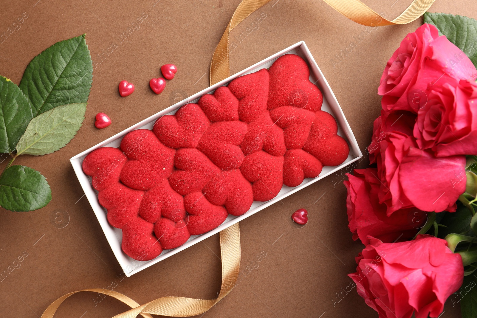 Photo of Chocolate bar decorated with hearts, confetti and roses on brown background, flat lay