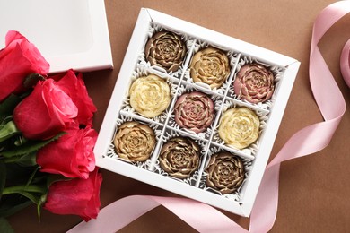 Photo of Delicious flower shaped chocolate bonbons in box and roses on brown background, flat lay