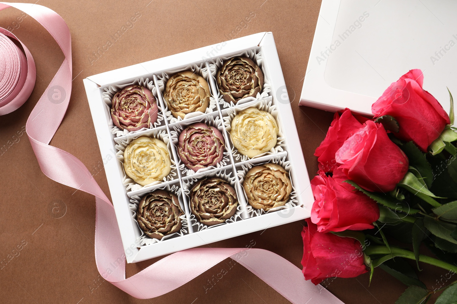 Photo of Delicious flower shaped chocolate bonbons in box and roses on brown background, flat lay