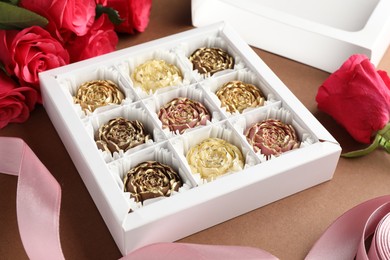 Photo of Delicious flower shaped chocolate bonbons in box and roses on brown background, closeup