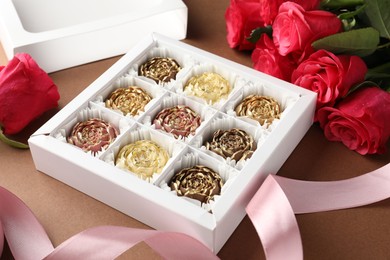 Photo of Delicious flower shaped chocolate bonbons in box and roses on brown background, closeup