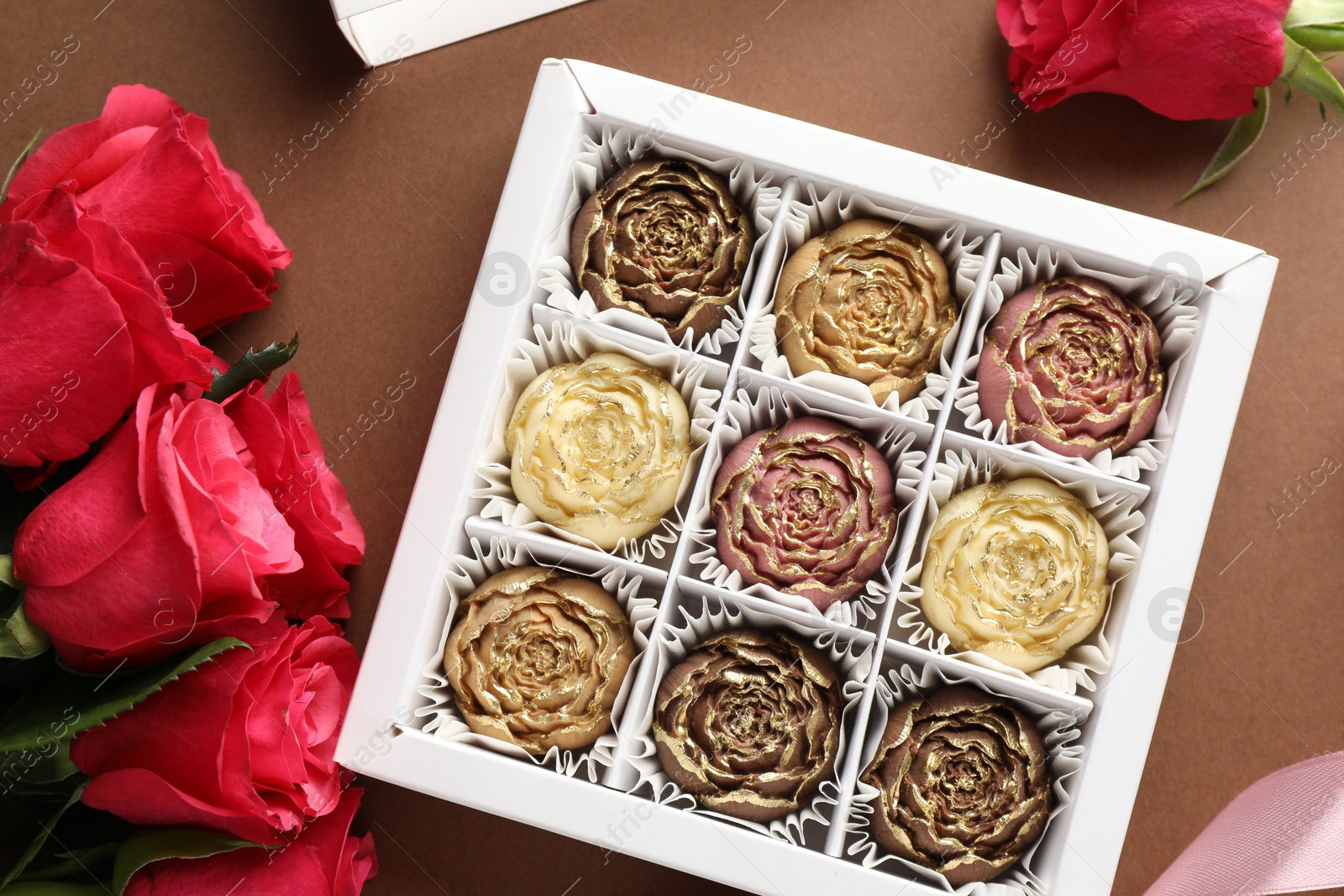 Photo of Delicious flower shaped chocolate bonbons in box and roses on brown background, flat lay