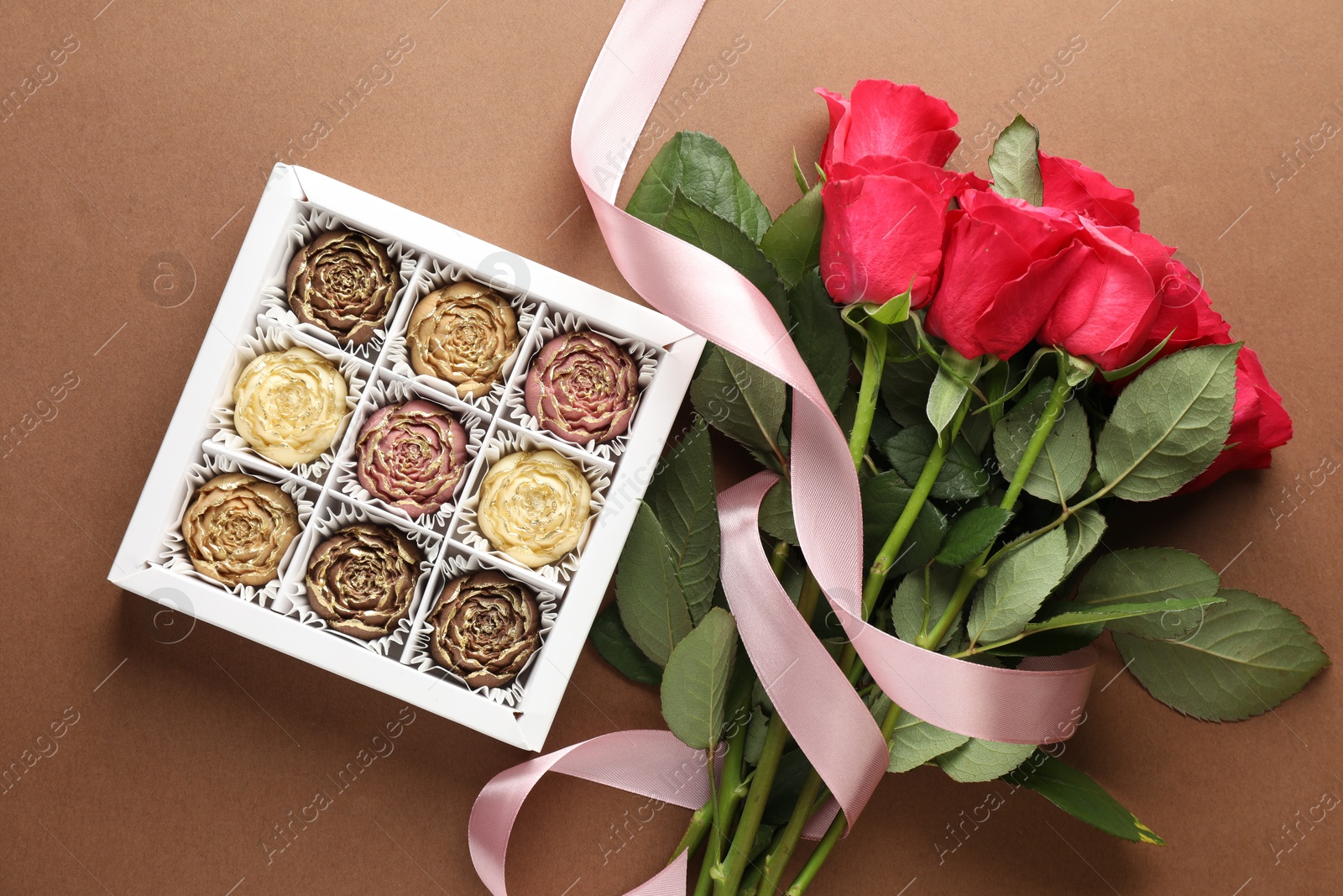 Photo of Delicious flower shaped chocolate bonbons in box and roses on brown background, flat lay