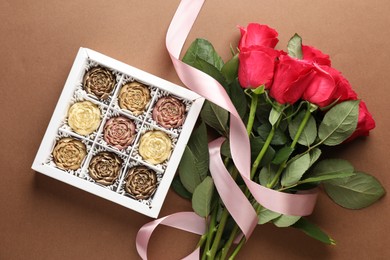 Photo of Delicious flower shaped chocolate bonbons in box and roses on brown background, flat lay