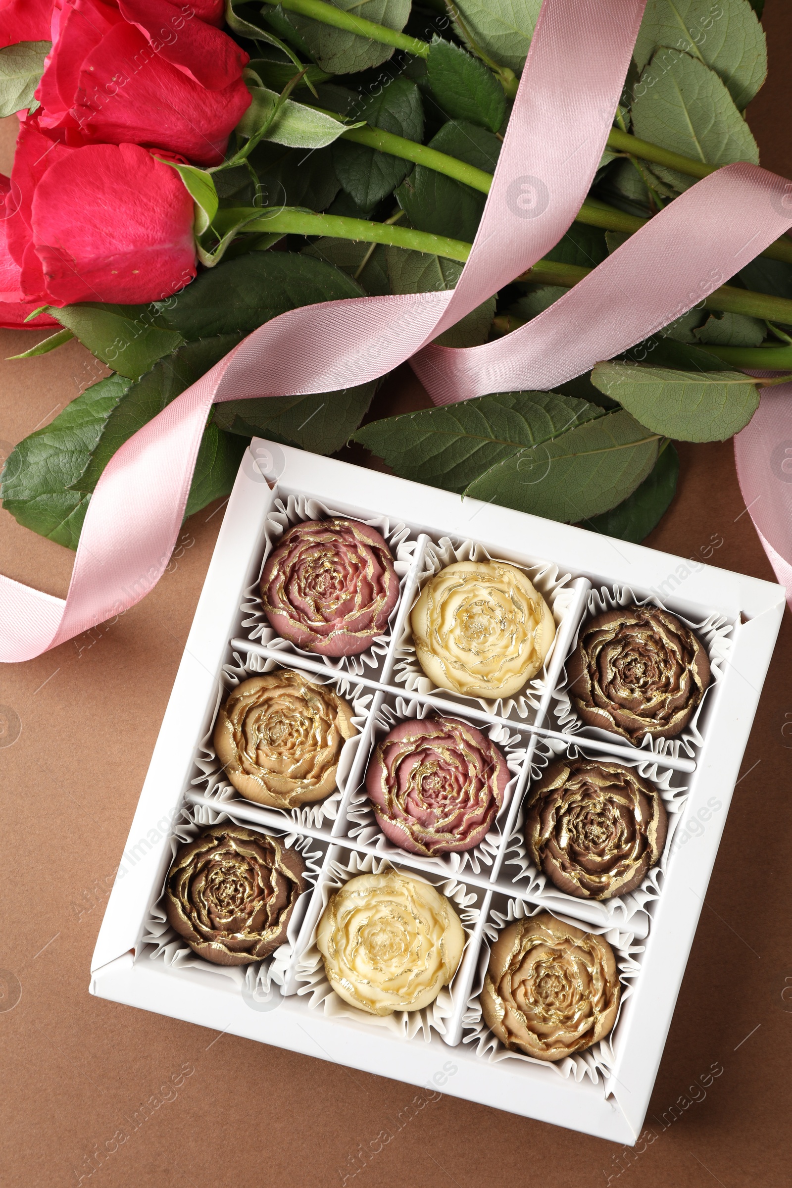 Photo of Delicious flower shaped chocolate bonbons in box and roses on brown background, flat lay