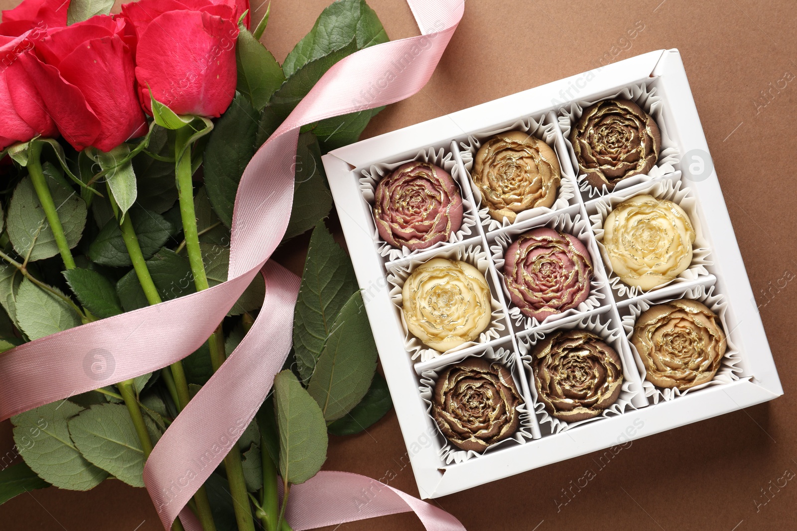 Photo of Delicious flower shaped chocolate bonbons in box and roses on brown background, flat lay