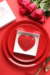 Photo of Heart shaped chocolate in box on plate and roses against red background, flat lay