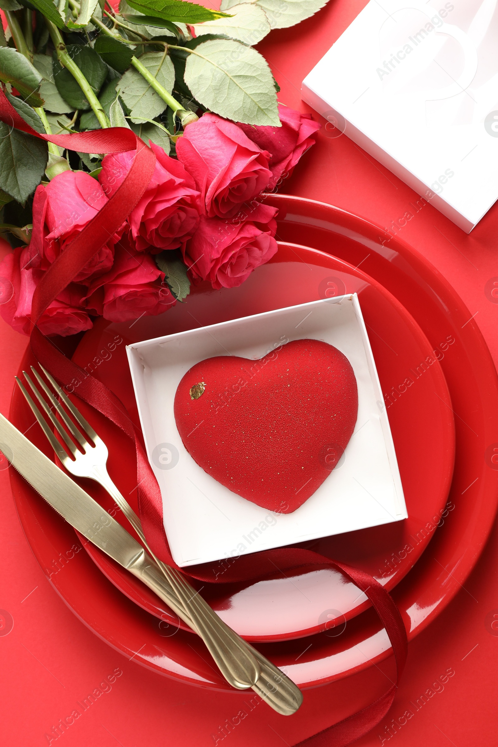 Photo of Heart shaped chocolate in box on plate and roses against red background, flat lay