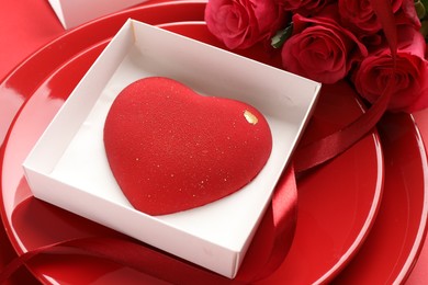 Photo of Heart shaped chocolate in box on plate and roses against red background, closeup