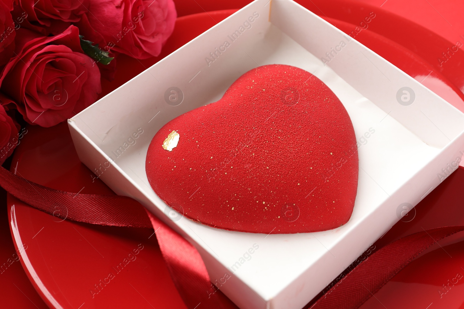 Photo of Heart shaped chocolate in box on plate and roses against red background, closeup