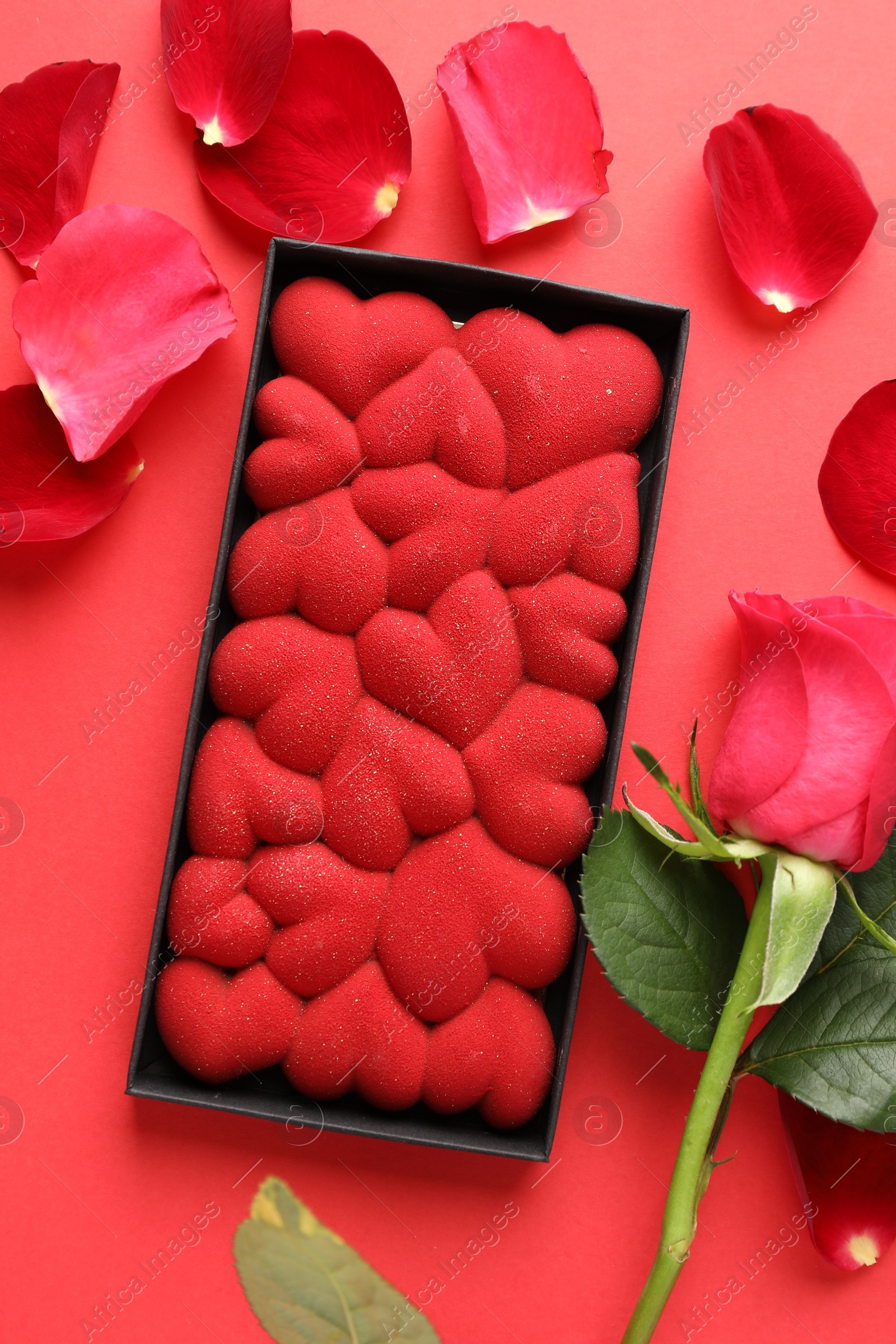 Photo of Chocolate bar decorated with hearts, rose and petals on red background, flat lay