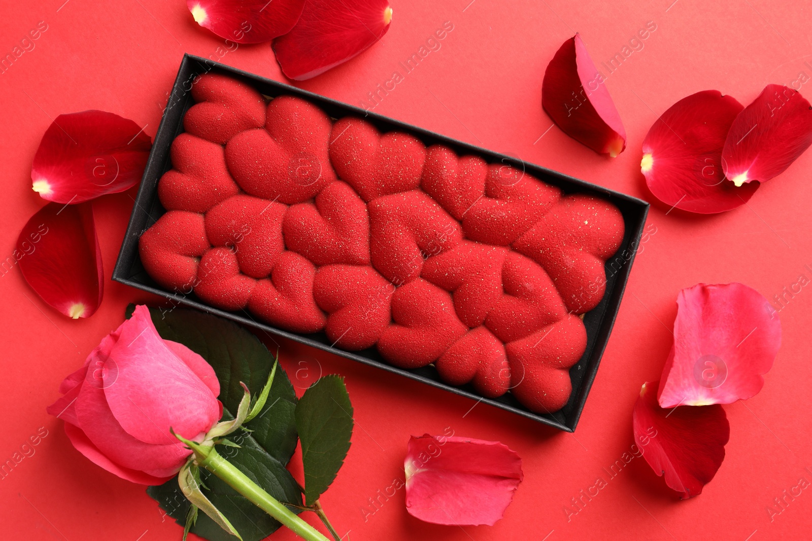 Photo of Chocolate bar decorated with hearts, rose and petals on red background, flat lay